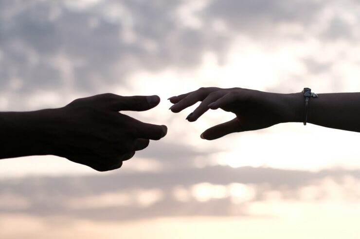 two peoples' hands reaching for each other, silhouetted against a cloudy sky