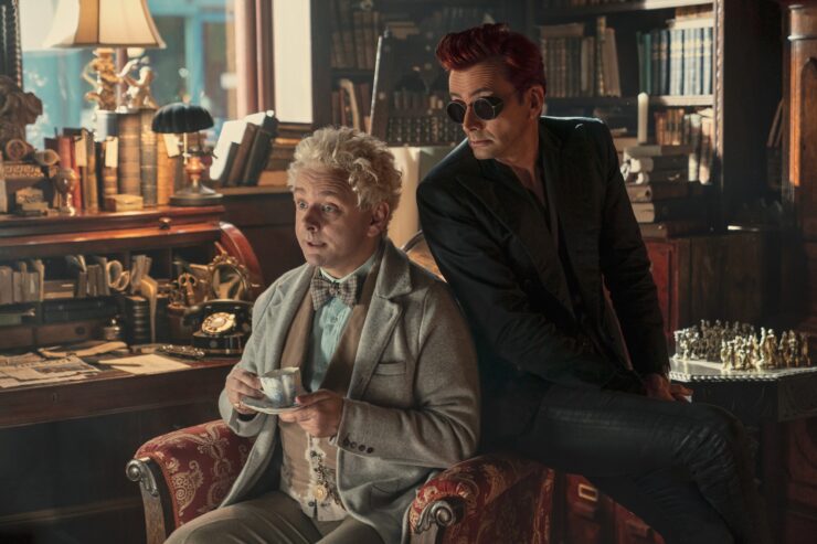 Good Omens 2, Aziraphale sitting in a chair in bookshop with tea, Crowley sitting on the arm directly behind him, both listening intently