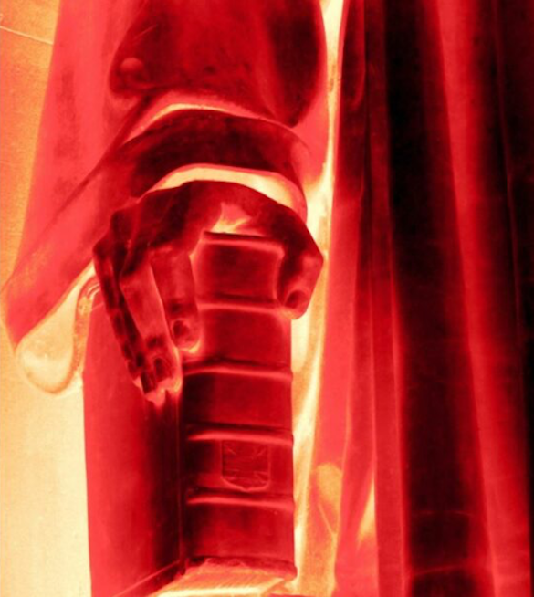 Photograph showing a close-up statue: the hand of a robed figure grasps a book. The photo has been manipulated to a high-contrast of reds, yellows, and oranges.