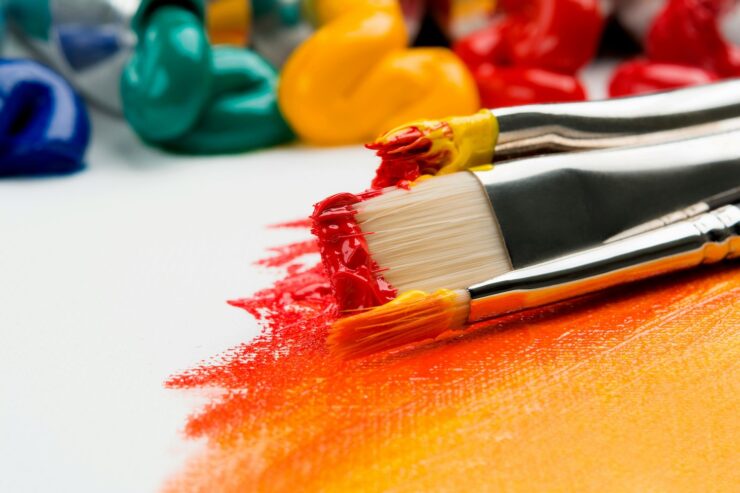 Close-up photograph of paint brushes covered in red, yellow, and orange paint, resting on a partially painted canvas. Paint tubes with dabs of blue, green, yellow, orange, and red paint appear in the background.