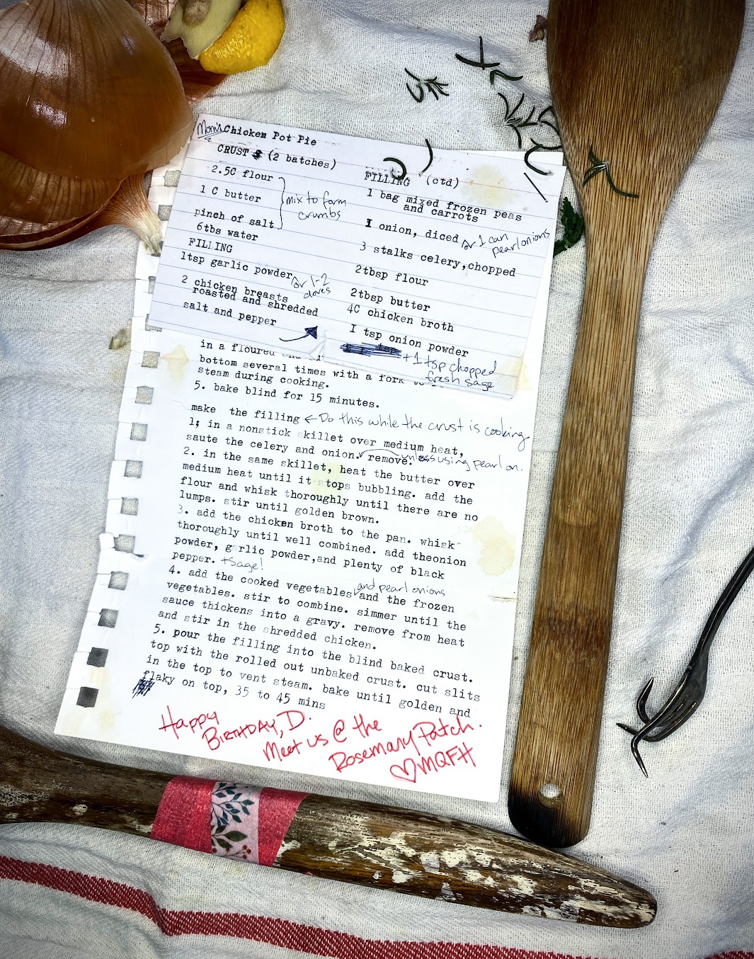 A recipe card, typewritten on an index card, stapled to a torn sheet of notebook paper with a typewritten recipe on it. Both are weathered, torn, stained, and annotated. The card is on top of a stained white kitchen towel, next to a couple of burned and repaired wooden cooking spoons, some onion and lemon scraps, a scattering of rosemary, and a bent fork. Visible recipe text is as follows (all is typewritten unless otherwise indicated; see story text for recipe in full):Index card: Chicken Pot Pie (handwritten annotation reading “Mom’s”)Crust (2 batches)2½ cups flour    Pinch of salt    1 cup butter (handwritten annotation indicates these three ingredients should be mixed to form crumbs)6 tablespoons waterFilling1 teaspoon garlic powder – handwritten annotation reading “or 1-2 cloves”2 chicken breasts roasted and shreddedSalt and pepper – hand-drawn arrow points up to the next column Filling continued1 bag mixed frozen peas and carrots1 onion, diced; handwritten annotation reading “or 1 can pearl onions”3 stalks celery, chopped2 tablespoons flour2 tablespoons butter4 cups chicken broth1 teaspoon onion powder(Several typing errors are scribbled out; handwritten annotation reading “1 tsp chopped fresh sage”)The index card overlaps the recipe page. The recipe visible on the page is as follows:5.	Bake blind for 15 minutes.  Make the Filling (handwritten annotation indicates to do this while the crust is cooking.)1.	In a nonstick skillet over medium heat, sauté the celery and onion. Handwritten note says “unless using pearl on.” Remove.2.	In the same skillet, heat the butter over medium heat until it stops bubbling. Add the flour and whisk thoroughly until there are no lumps. Stir until golden brown.3.	Add the chicken broth to the pan. Whisk thoroughly until well-combined. Add the onion powder, garlic powder, and plenty of black pepper. (Handwritten annotation reads “+ sage!”)4.	Add the cooked vegetables (handwritten annotation reads “^or 1 can pearl onions”) and the frozen vegetables. Stir to combine. Simmer until the sauce thickens into a gravy. Remove from the heat and stir in the shredded chicken. 7.	Pour the filling into the blind-baked crust. Top with the rolled-out unbaked crust. Cut slits in the top to vent steam. Bake until golden and flaky on top, 35–45 minutes. Below the recipe, a typing error is scribbled out. A handwritten note in different handwriting from the recipe annotations, in red marker, reads: “Happy birthday, D. Meet us @ the Rosemary Patch. Heart, MQFH”