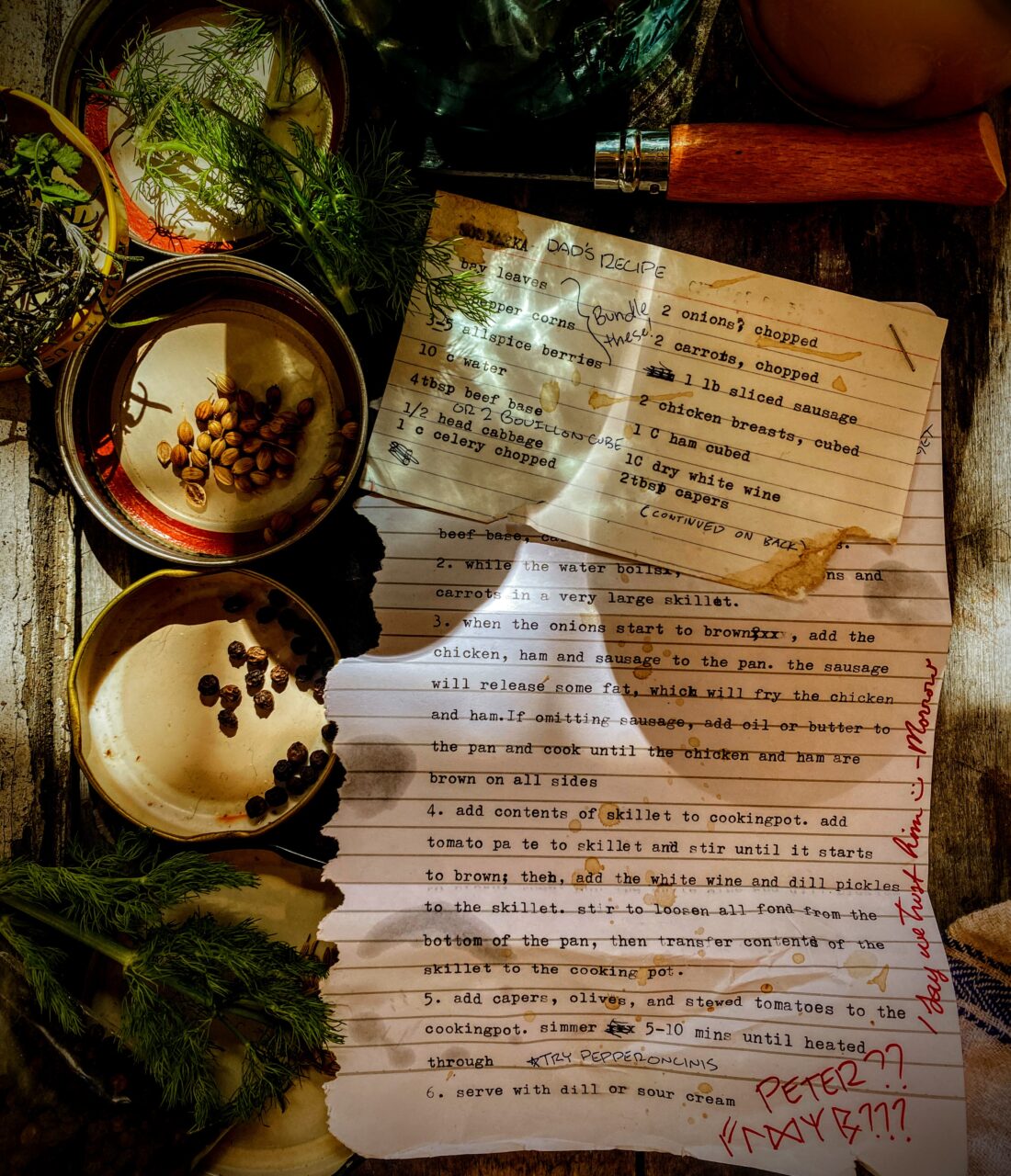 A recipe card, typewritten on an index card, stapled to a torn sheet of notebook paper with a typewritten recipe on it. Both are weathered, torn, stained, and annotated. The card is on top of weathered, scarred wood, and is surrounded by jar lids holding whole spices, dill fronds, a folding knife, and a couple of jars with preserved vegetables and meats.

Visible recipe text is as follows (all is typewritten unless otherwise indicated; see story text for recipe in full):

The name of this recipe is blurred out from damage to the recipe card. Handwritten annotation says “Dad’s Recipe.”
2 bay leaves
peppercorns
3–5 allspice berries (handwritten annotation indicates to bundle these ingredients)
10 c water
4 tablespoons beef base *handwritten annotation suggests substituting 2 bouillon cubes)
½ head cabbage
1 c celery, chopped
2 onions, chopped
2 carrots, chopped
1 pound sliced sausage
2 chicken breasts, cubed
1 cup ham, cubed
1 cup dry white wine
2 tablespoons capers
(Handwritten annotation reads “continued on back.”)
The index card overlaps the recipe page. The recipe visible on the page is as follows:
2. While the water boils [obscured] and carrots in a very large skillet.
3. When the onions start to brown, add the sausage, chicken, and ham to the pan. The sausage will release some fat, which will fry the chicken and ham. If omitting sausage, add oil or butter to the pan and cook until the chicken and ham are brown on all sides.
4. Add the contents of the skillet to the cooking pot. Add tomato paste to the skillet and stir until it starts to brown; then, add the white wine and dill pickles to the skillet. Stir to loosen all fond from the bottom of the pan, then transfer contents of the skillet to the cooking pot.
5. Add capers, olives, and stewed tomatoes to the cooking pot. Simmer 5–10 minutes until heated through. (Handwritten annotation reads “*try pepperoncinis”)
6. Serve with dill or sour cream

A handwritten note in different handwriting from the recipe annotations, in red marker, reads: “Peter??” followed by a series of runes. Next to that, running up the side of the page, a handwritten note in sweet cursive, also in red marker, reads “I say we trust him :) -morrow”
