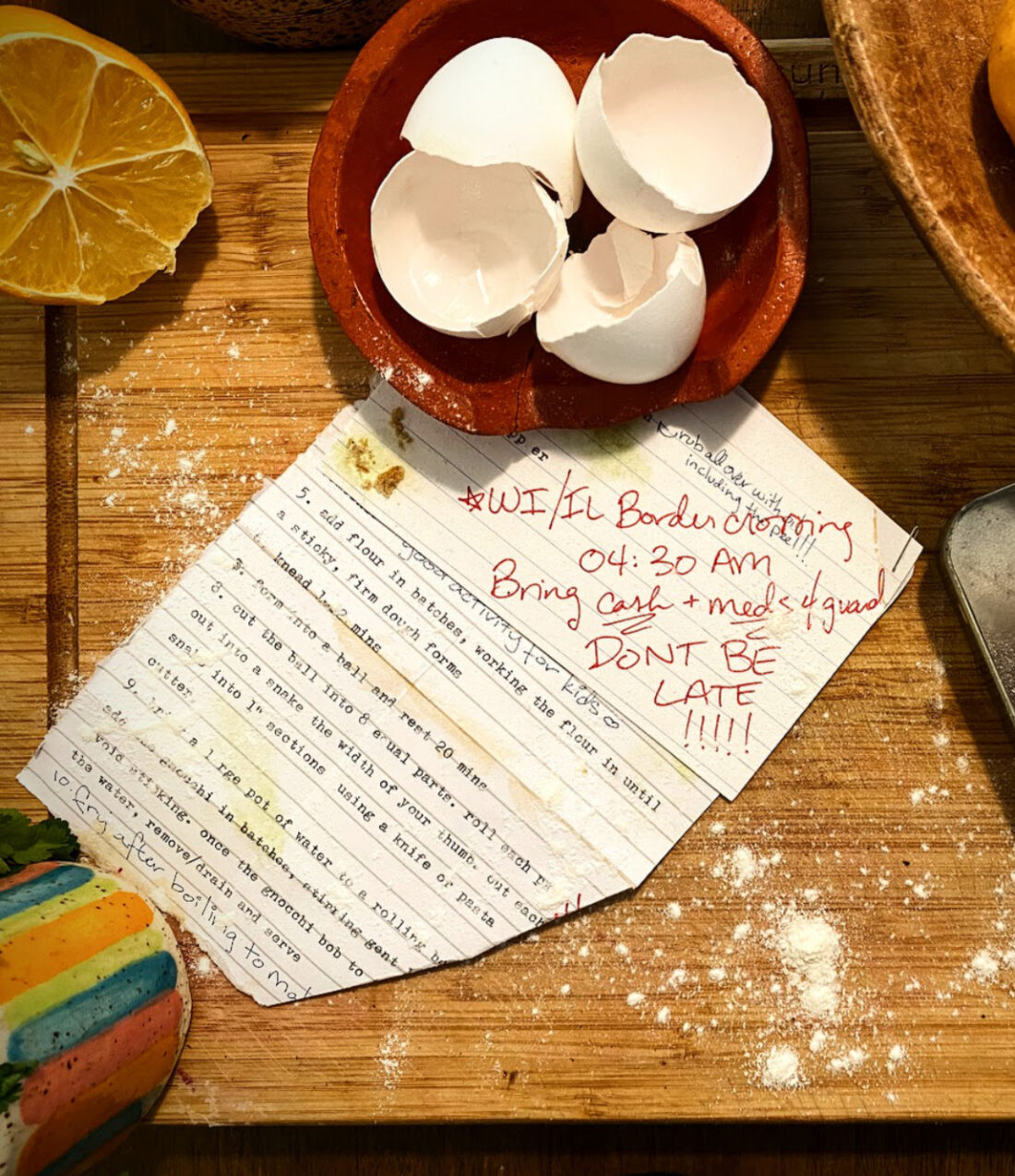 A recipe card, typewritten on an index card, stapled to a torn sheet of notebook paper with a typewritten recipe on it. Both are weathered, torn, stained, and annotated. The card is on top of a hefty wooden cutting board, and is surrounded by a bowl of lemons, a dish of eggshells, a cut lemon, a repurposed breath mints tin containing mixed pills including estrogen/estradiol and antidepressants, and scattered flour. Much of the recipe card is obscured by the eggshells. 

Visible recipe text is as follows (all is typewritten unless otherwise indicated; see story text for recipe in full): 

On the recipe card: a handwritten note reading ‘rub all over with oil including the peel!!!’ is overlapped by a handwritten note in different handwriting, in red marker, which reads: “WI / IL border crossing 04:30 AM Bring CASH and MEDS 4 guard don’t be late!!!!!!!”
The index card overlaps the recipe page. The recipe visible on the page is as follows:

5.	Add flour in batches, working the flour in until a sticky, firm dough forms. 
6.	Knead 1–2 mins
7.	Form into a ball and rest for 20 mins
8.	Cut the ball into 8 equal parts. Roll each part out into a snake the width of your thumb. Cut each snake into 1-inch sections using a knife or pasta cutter.
9.	[after this point, the recipe card is folded and obstructs part of each line.] Bring a large pot of water to a rolling b. Add the gnocchi in batches, stirring gent … void sticking. Once the gnocchi bob … the water, remove/drain and serve.
Partially obscured handwritten annotation reads: Fry after boiling to [text obscured]
