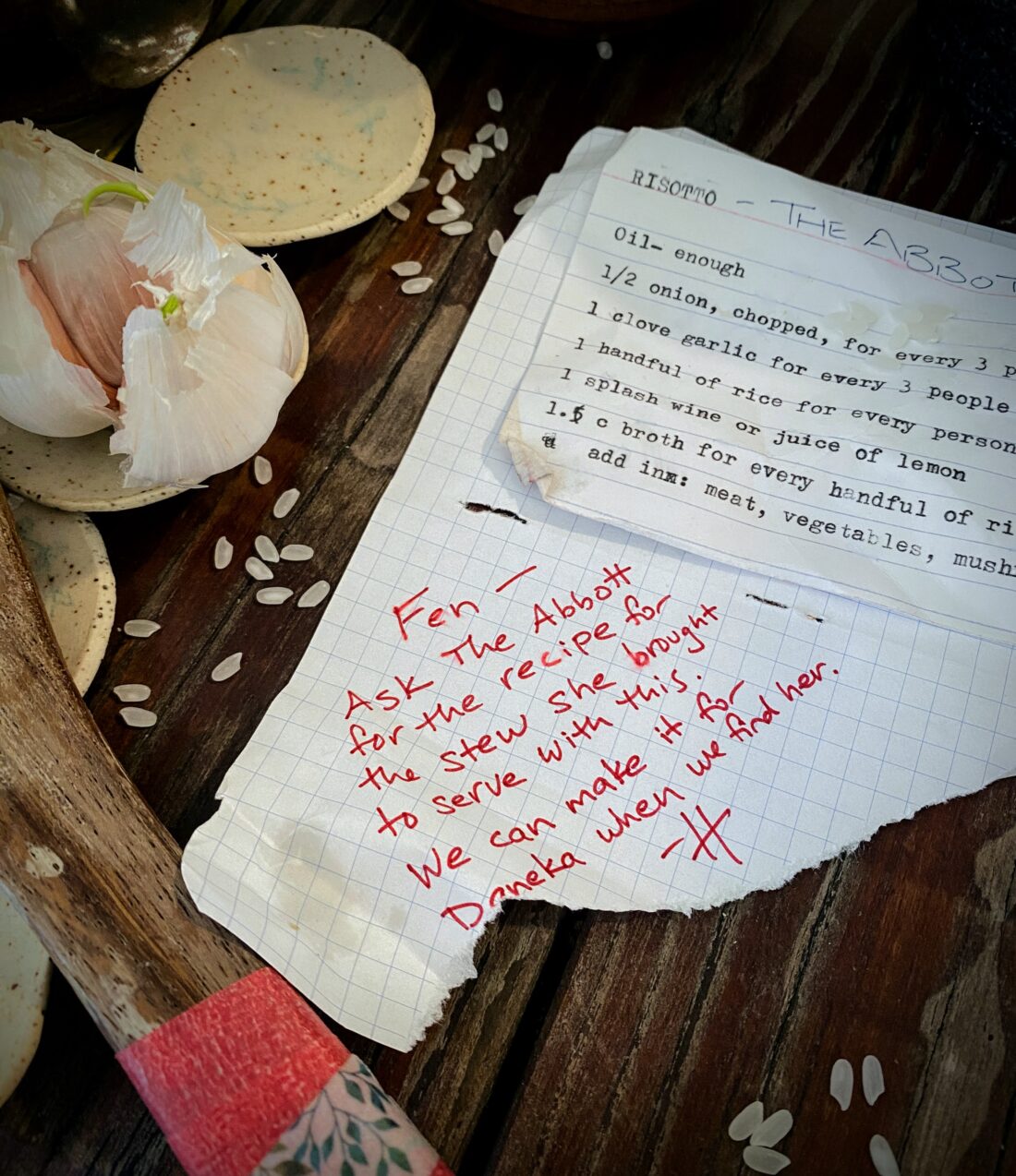 A recipe card, typewritten on an index card, stapled to a torn sheet of notebook paper with a typewritten recipe on it. Both are weathered, torn, stained, and annotated. The card is on top of sturdy, well-worn wood, and is surrounded by a repaired wooden spoon, small dishes holding sprouted garlic, and some scattered short-grain rice. Visible recipe text is as follows (all is typewritten unless otherwise indicated. Recipe card cuts off at the edge of the image; see story text for recipe in full): On the recipe card: Risotto – a handwritten annotation adds “The Abbott” Oil—enough ½ onion, chopped, for every 3 1 clove garlic for every 3 people 1 handful of rice for every person 1 splash wine or juice of 1 lemon 1½ cups broth for every handful of ri Add in: meat, vegetables, mush The recipe page beneath does not have visible recipe text. A handwritten note in red marker, in a unique handwriting, says: “Fen – Ask The Abbott for the recipe for the stew she brought to serve with this. We can make it for Daneka when we find her. -H”