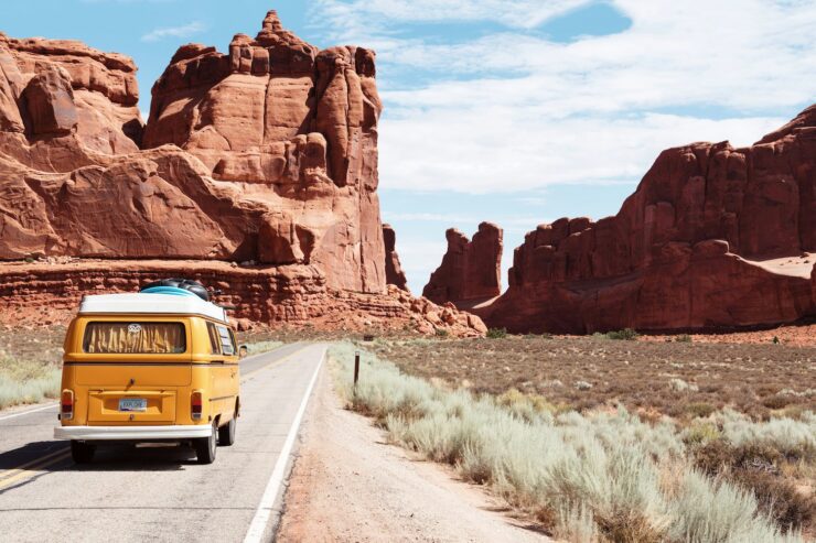 vintage yellow Volkswagen bus on a road, driving away from the camera. The andscape is a desert, with scrub brush at the side of the road and buttes in the background.