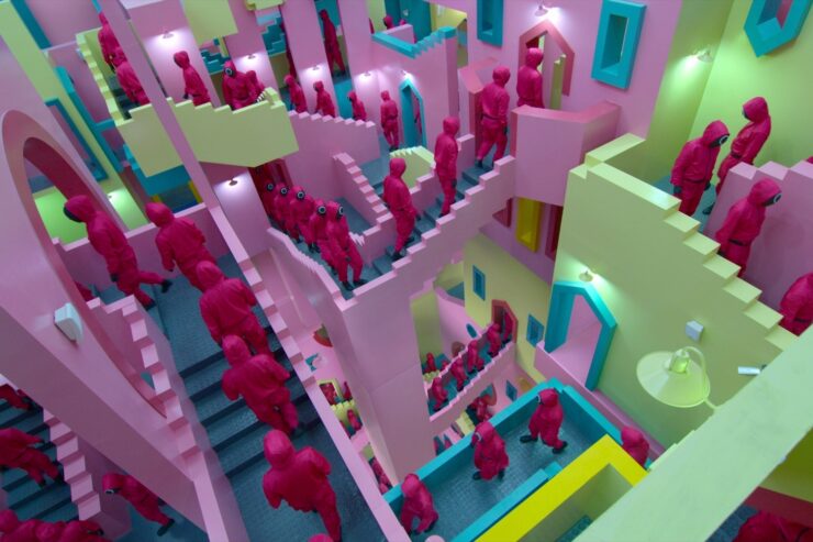 An image from the TV series Squid Game: masked figures walk single-file on a series of brightly-colored staircases