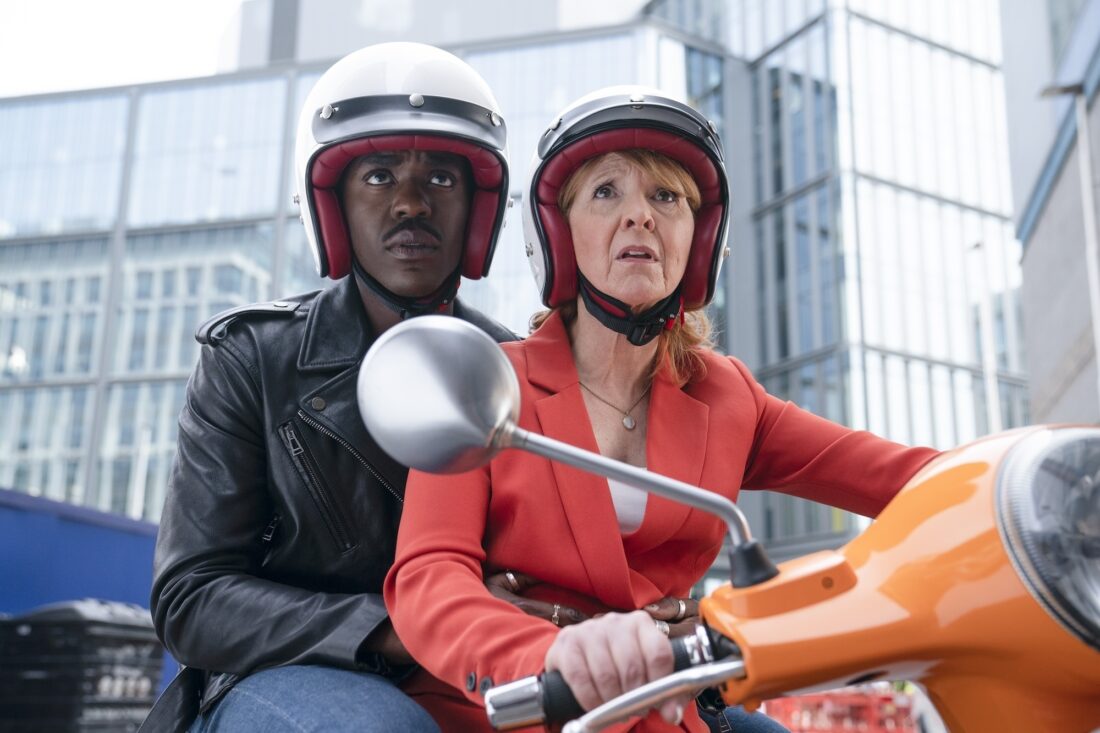 Melanie Bush (Bonnie Langford) and the Doctor (Ncuti Gatwa) on Mel's orange scooter on Doctor Who, Empire of Death