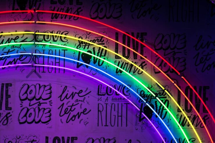 Photo of a neon light rainbow against a wall decorated with phrases including "Love is Love", "Support Love", and "Live and Let Love"