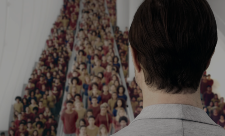 Scene from the TV series 3% (Três por cento): A male figure dressed in light grey in the foreground stands facing a large homogeneous crowd dressed in yellow, red, and blue t-shirts.