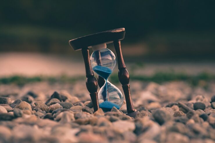 Photo of an hourglass with blue sand, resting at an angle among rocks.