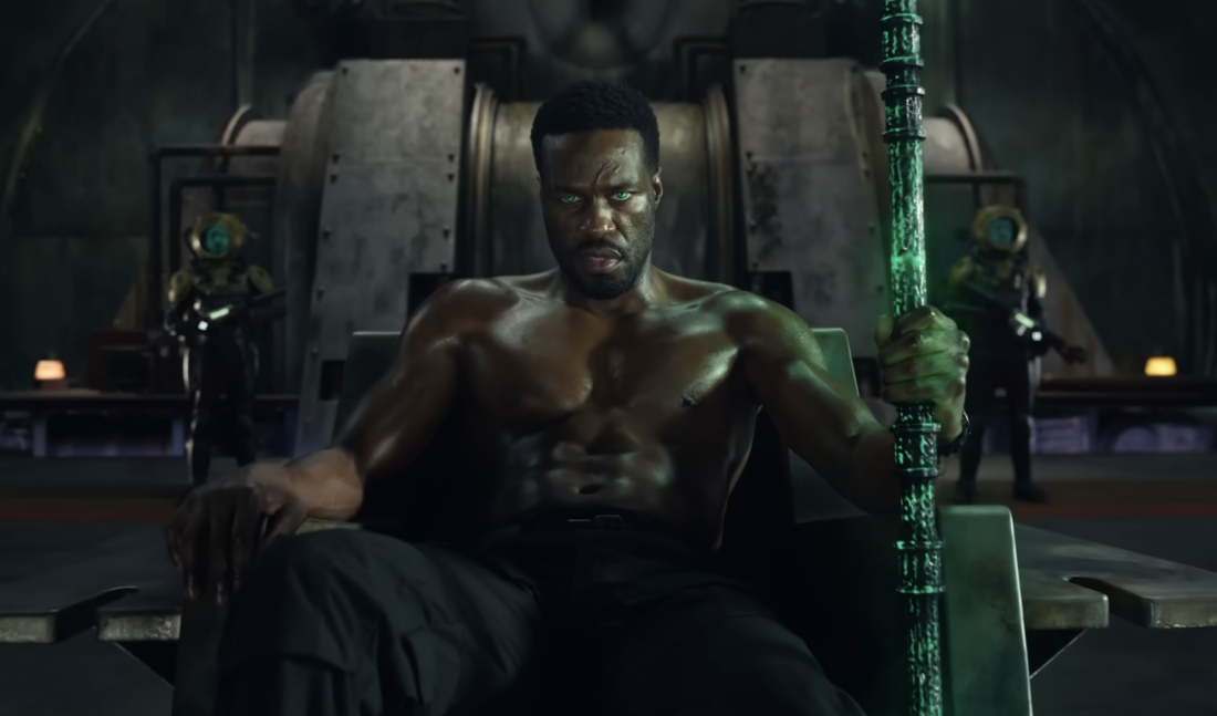 A scene from Aquaman and the Lost Kingdom: Black Manta (Yahya Abdul-Mateen II) sits on a metalic throne, holding a glowing green trident.