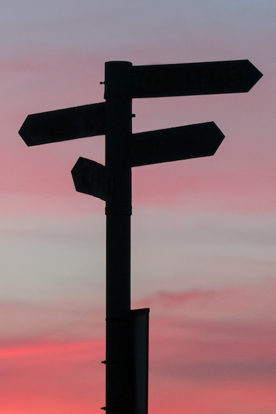 Photo of a road sign with directional arrows pointing in different directions, silhouetted against a sky at sunset