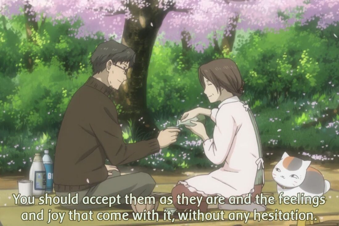 A scene from the anime series Natsume's Book of Friends: a woman pours a drink for a man in an outdoor setting with a cat sitting nearby. The subtitles read: "You should accept them as they are and the feelings and joy that come with it, without any hesitation"