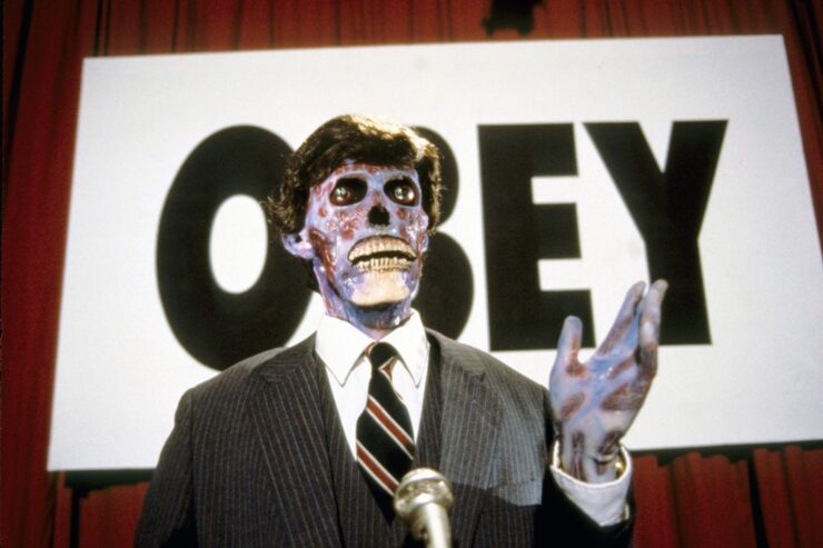 An alien disguised as a human stands in front of a sign declaring "OBEY" in a scene from They Live