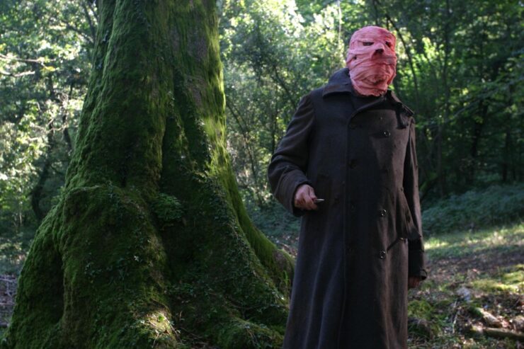 A figure in a long coat stands in the woods; their face is wrapped in bandages and they carry scissors. From the movie Timecrimes.