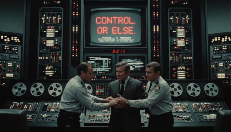 Image from the film Colossus: The Forbin Project. Two officers restrain a man in a suit in front of a large supercomputer. A screen in the center of the image reads "Control. Or Else."